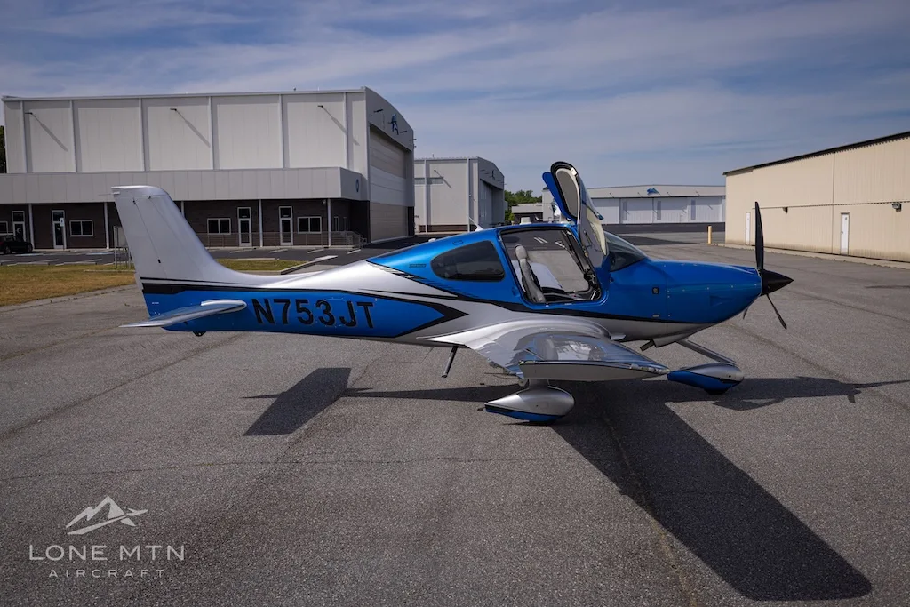 A blue and silver single-engine aircraft, N753JT, with open gull-wing doors is parked on an asphalt tarmac next to hangars.