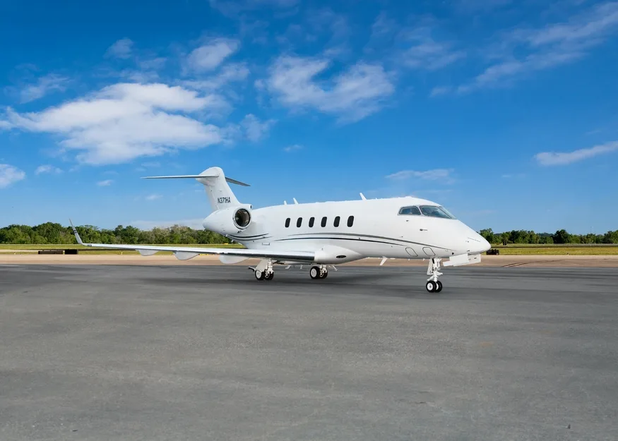 A white Bombardier Challenger business jet with a black stripe on the tarmac under a blue sky.