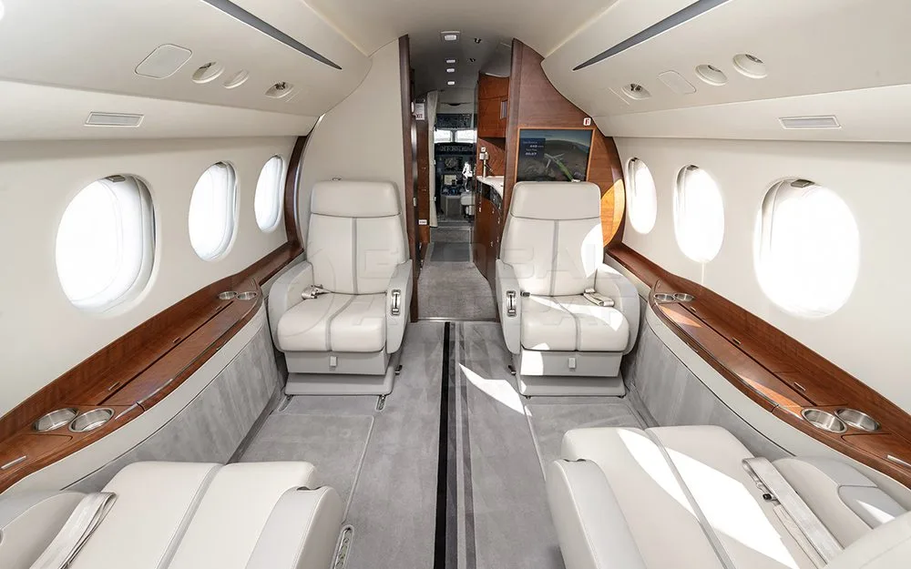 A view down the aisle of a private jet's cabin, showing rows of light-colored leather seats and a screen at the far end.