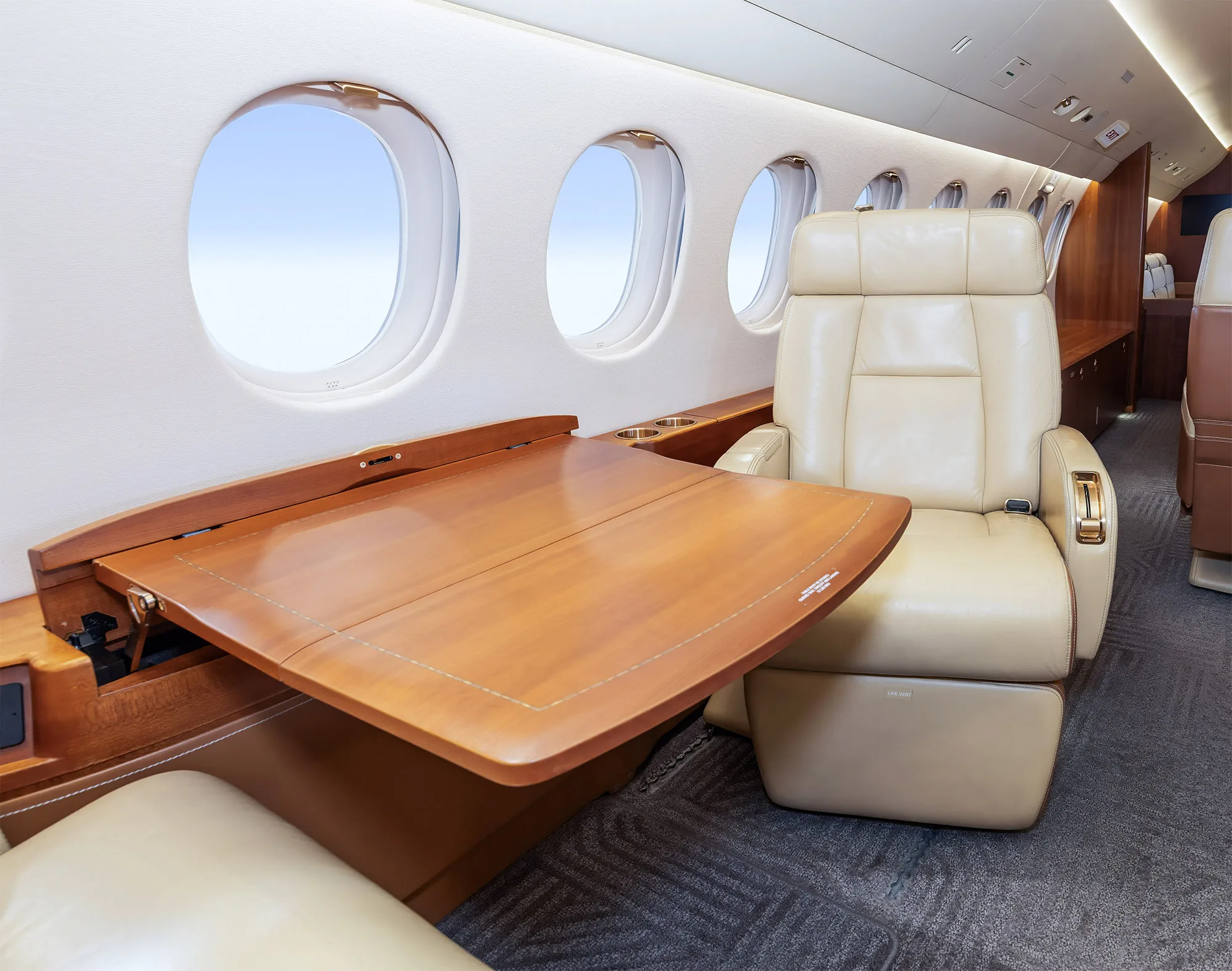 A close-up of the business jet's cabin interior, showing a beige leather seat and a large wooden table extended from the side panel.