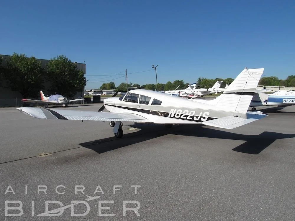 A rear-side view of the white Piper Arrow 200 (N822JS) with black and grey stripes, parked on an asphalt tarmac with other aircraft in the background.