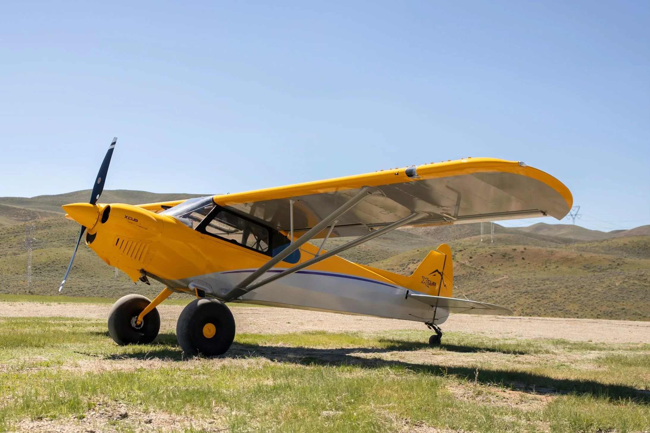 A yellow and silver CubCrafters XCub aircraft with large bush tires parked on a grassy field under a clear blue sky.