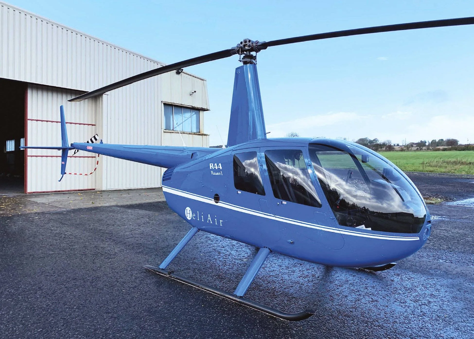 A blue Robinson R44 helicopter with "Heli Air" branding parked on a wet tarmac in front of a hangar.