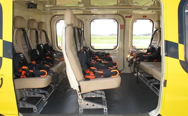 The interior cabin of a helicopter showing multiple beige seats with orange safety harnesses.
