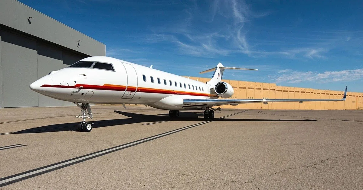 A white Bombardier Global business jet with red and orange stripes on the fuselage is parked on an airfield.