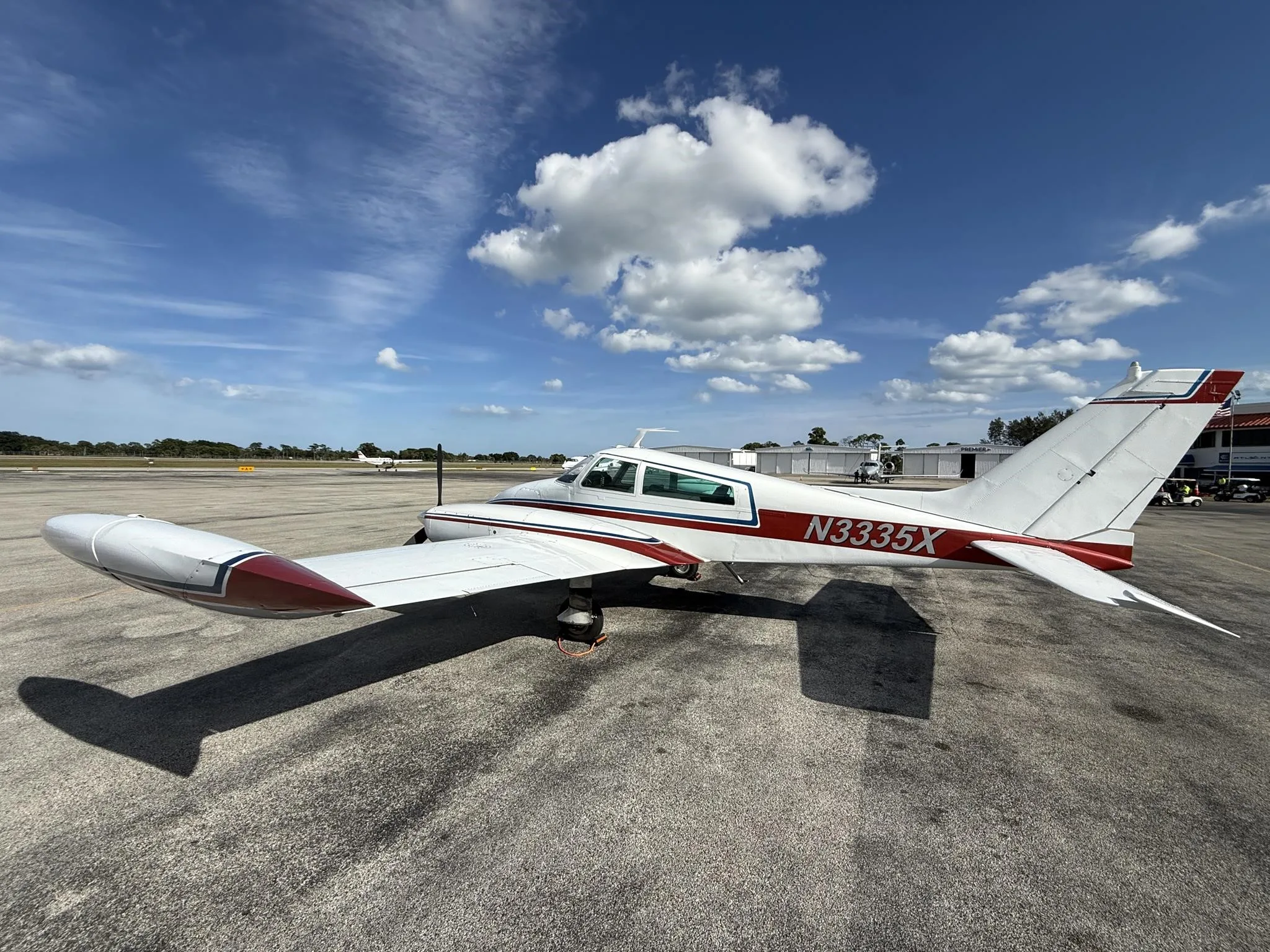 A white Cessna 310L with red and blue stripes parked on an airport tarmac under a partly cloudy sky.