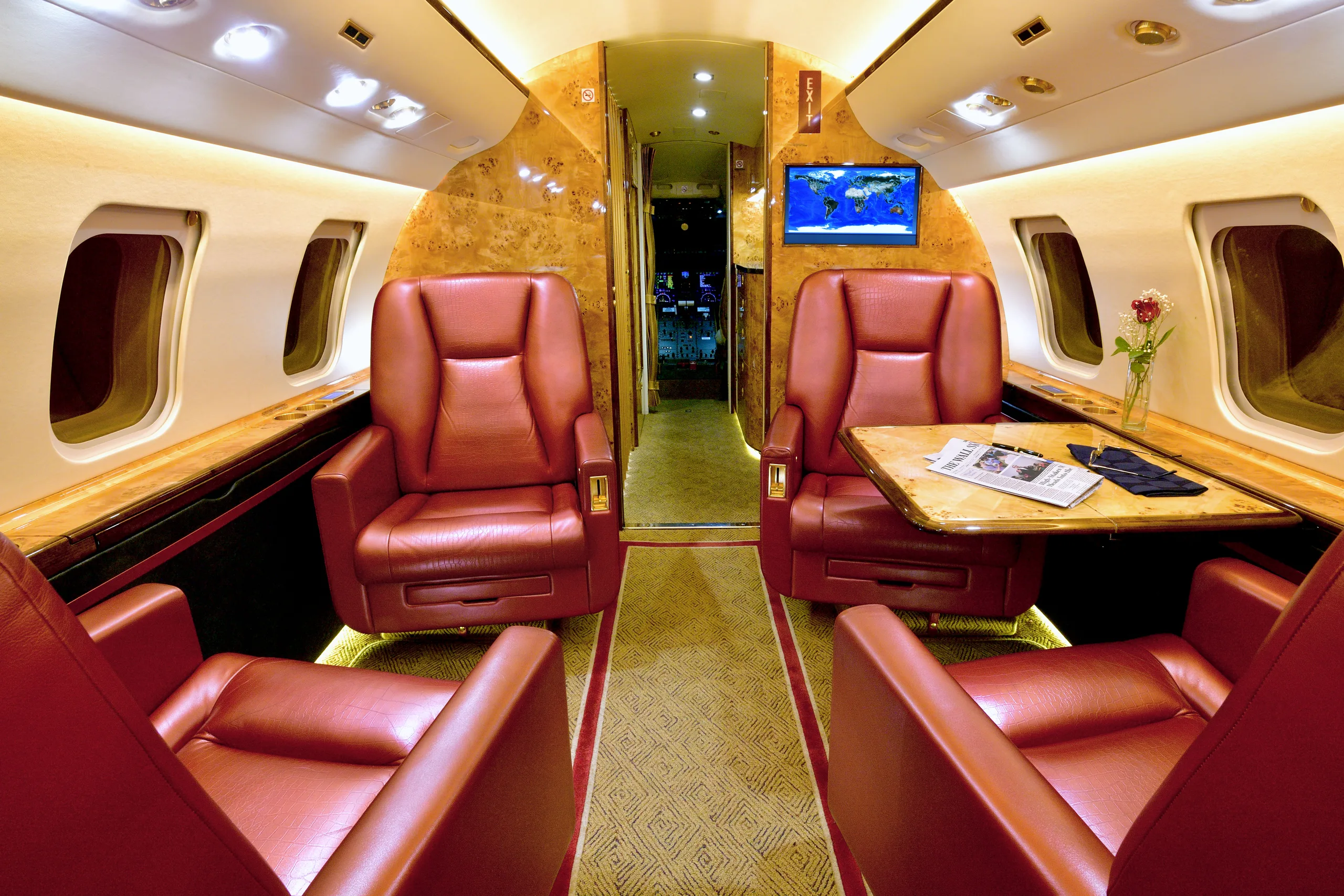 The elegant cabin interior of a private jet featuring red leather seats, polished wood accents, and a screen displaying a map.