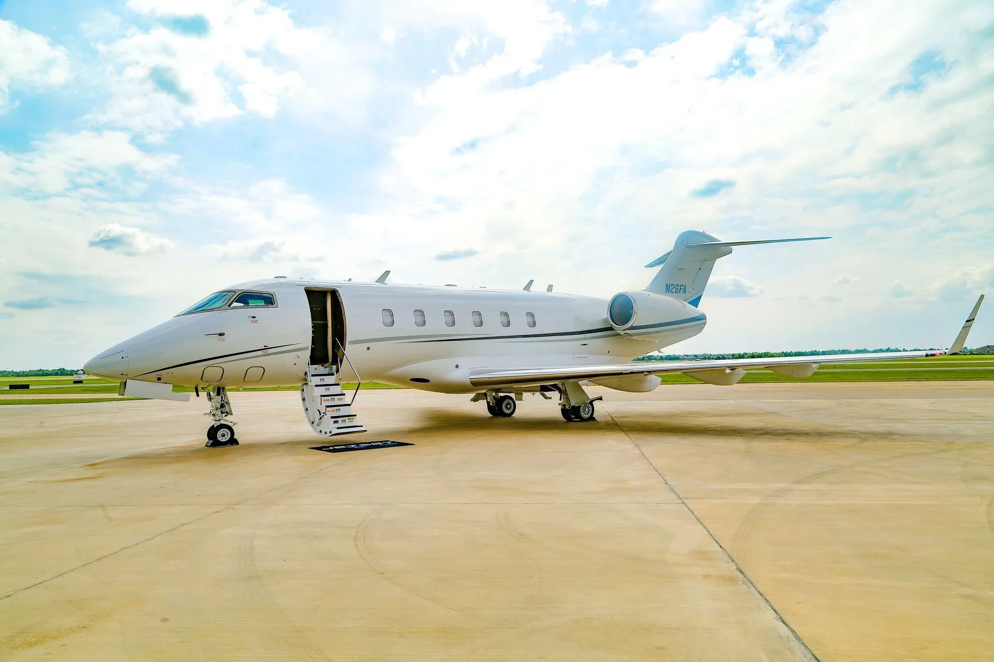 A white Bombardier Challenger 350 business jet, registration N26FA, is parked on a tarmac with its main door open and stairs extended.