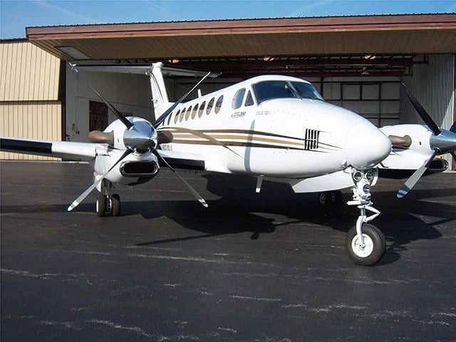 A white Beechcraft King Air with brown and black stripes parked outside a hangar with its door open.