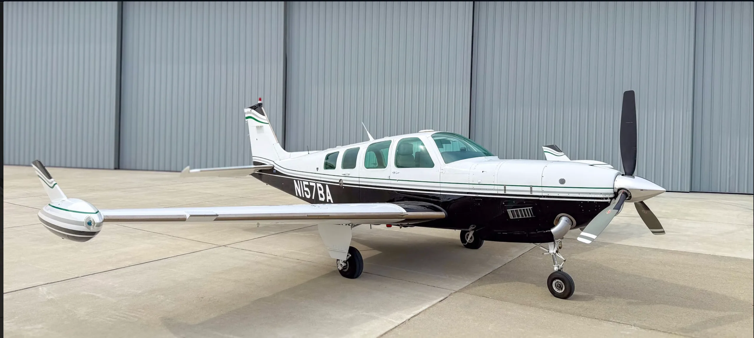 A white and black Beechcraft Baron N157BA with green stripes, parked on an airfield in front of a hangar.