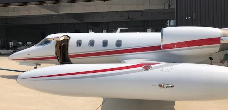 A close-up side view of a white Learjet with red and grey stripes, showing the open passenger door.