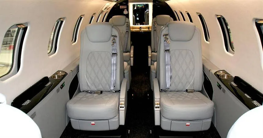 The interior cabin of a private jet with light grey quilted leather seats and windows.