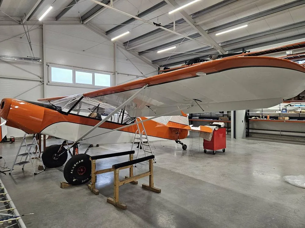 An orange and white high-wing aircraft with large bush tires, parked in a hangar with ladders nearby.