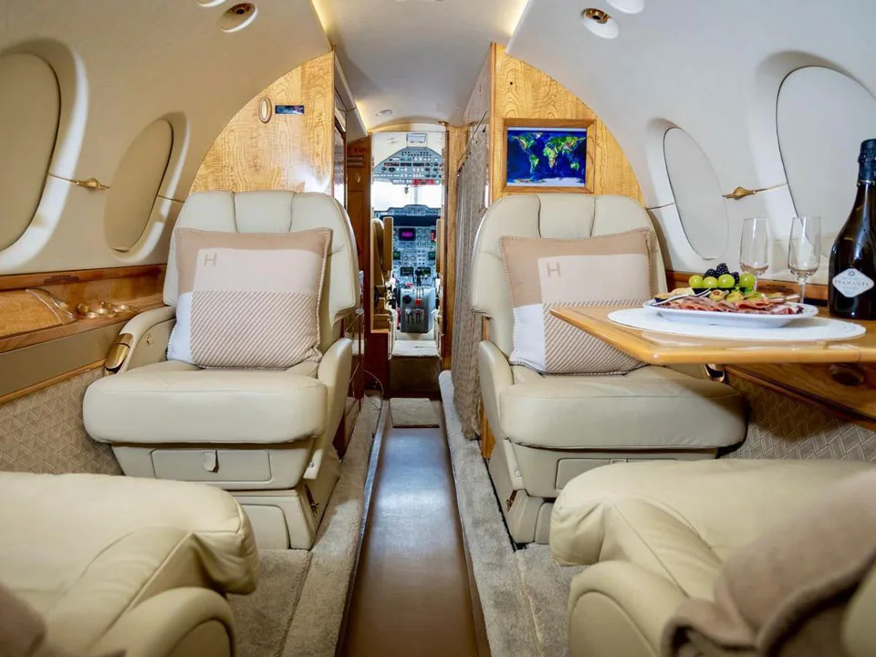 A view down the aisle of a private jet's luxurious cabin, showing cream leather seats, wood paneling, and a table set with refreshments.