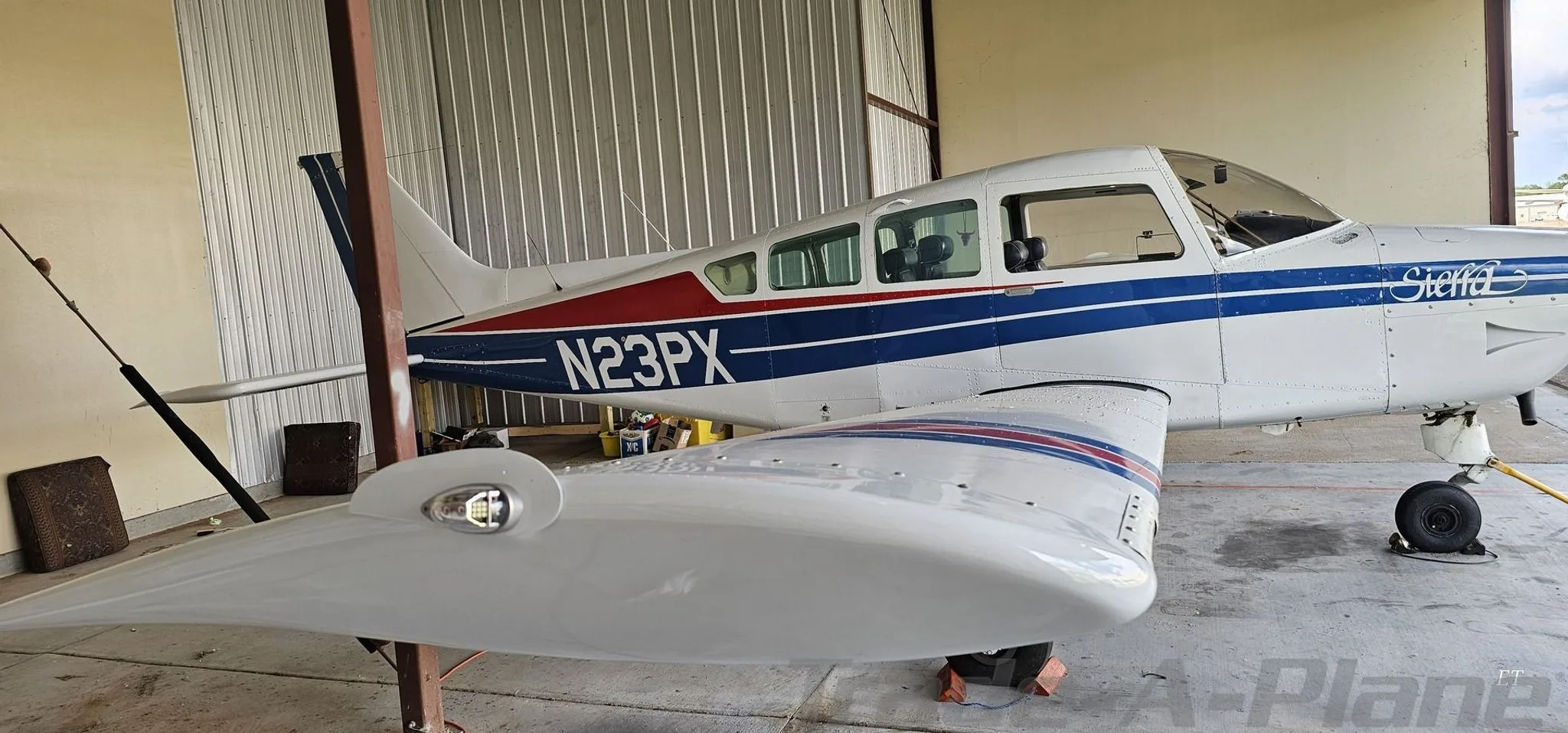 A white Beechcraft Sierra N23PX with blue and red stripes parked in a hangar, showing the right wing and fuselage.