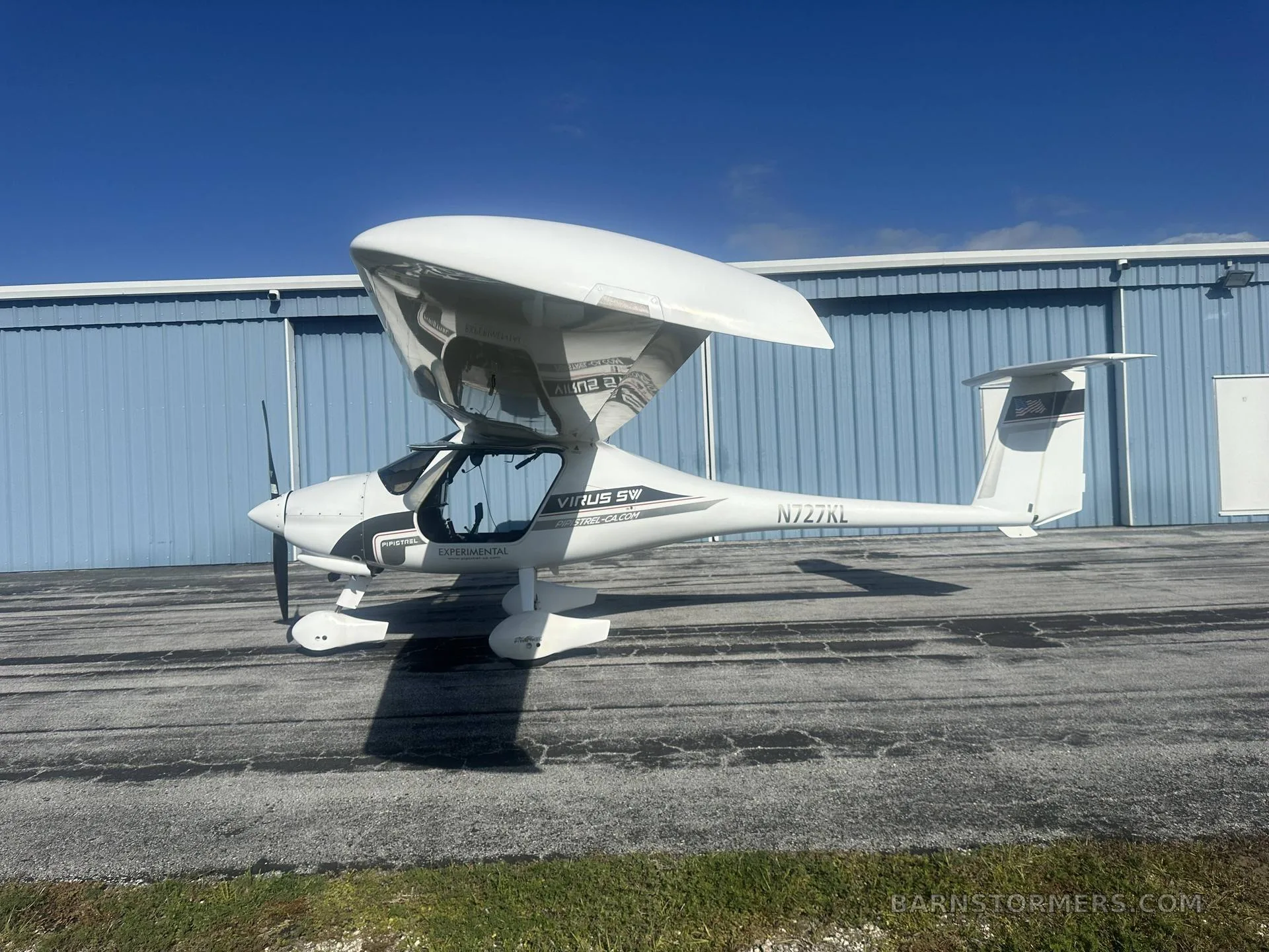 A white Pipistrel Virus SW aircraft with its wings folded up is parked on a tarmac next to a blue hangar.