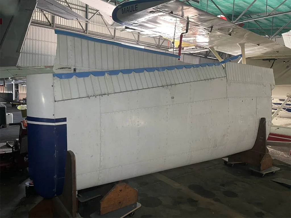 The white fuselage of an aircraft with a blue stripe is supported on wooden stands inside a hangar.