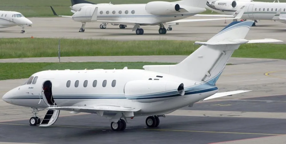 A white business jet with blue stripes is parked on an airport tarmac with its main door open and stairs extended.