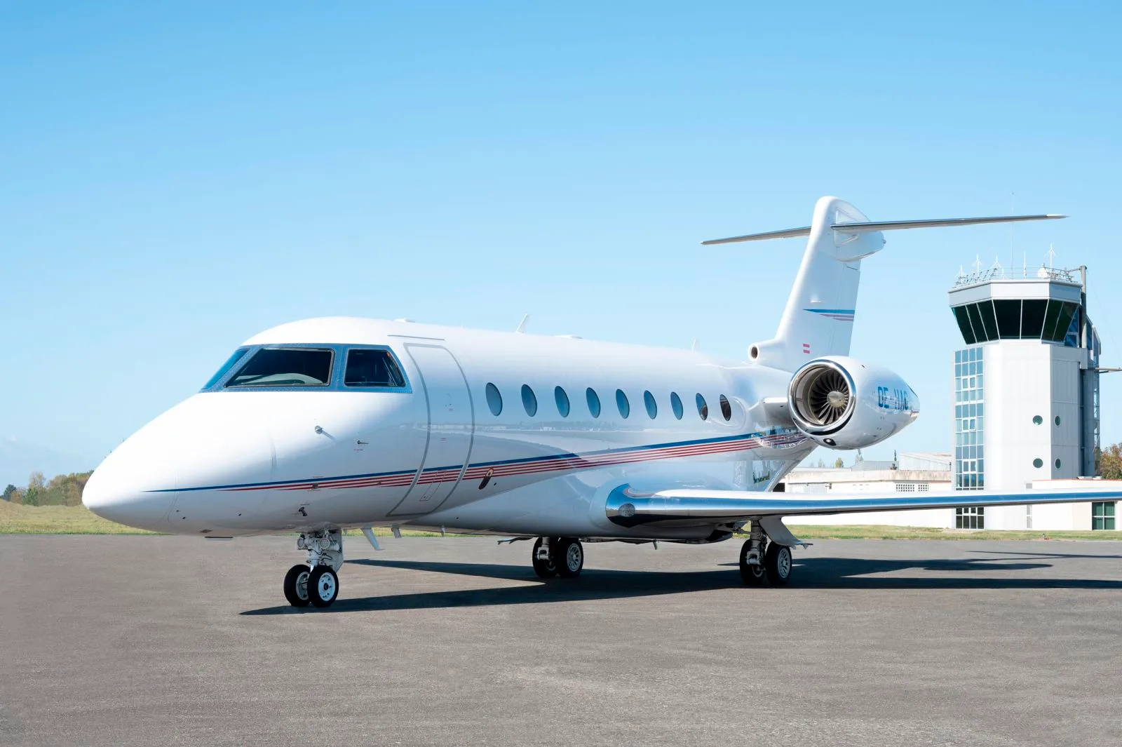A white business jet with red and blue stripes is parked on an airport tarmac, with an air traffic control tower in the background.