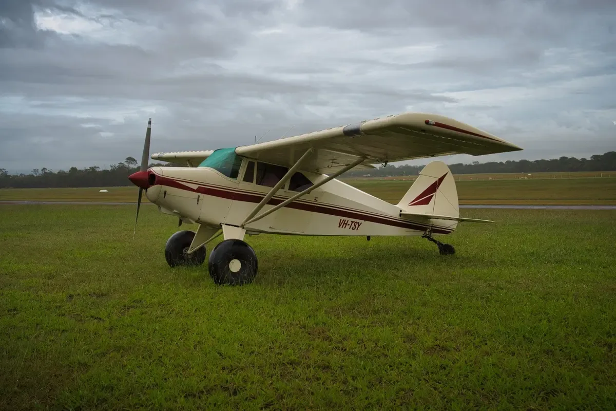A cream and maroon single-engine taildragger aircraft with registration VH-TSY parked on a grassy airfield under a cloudy sky.