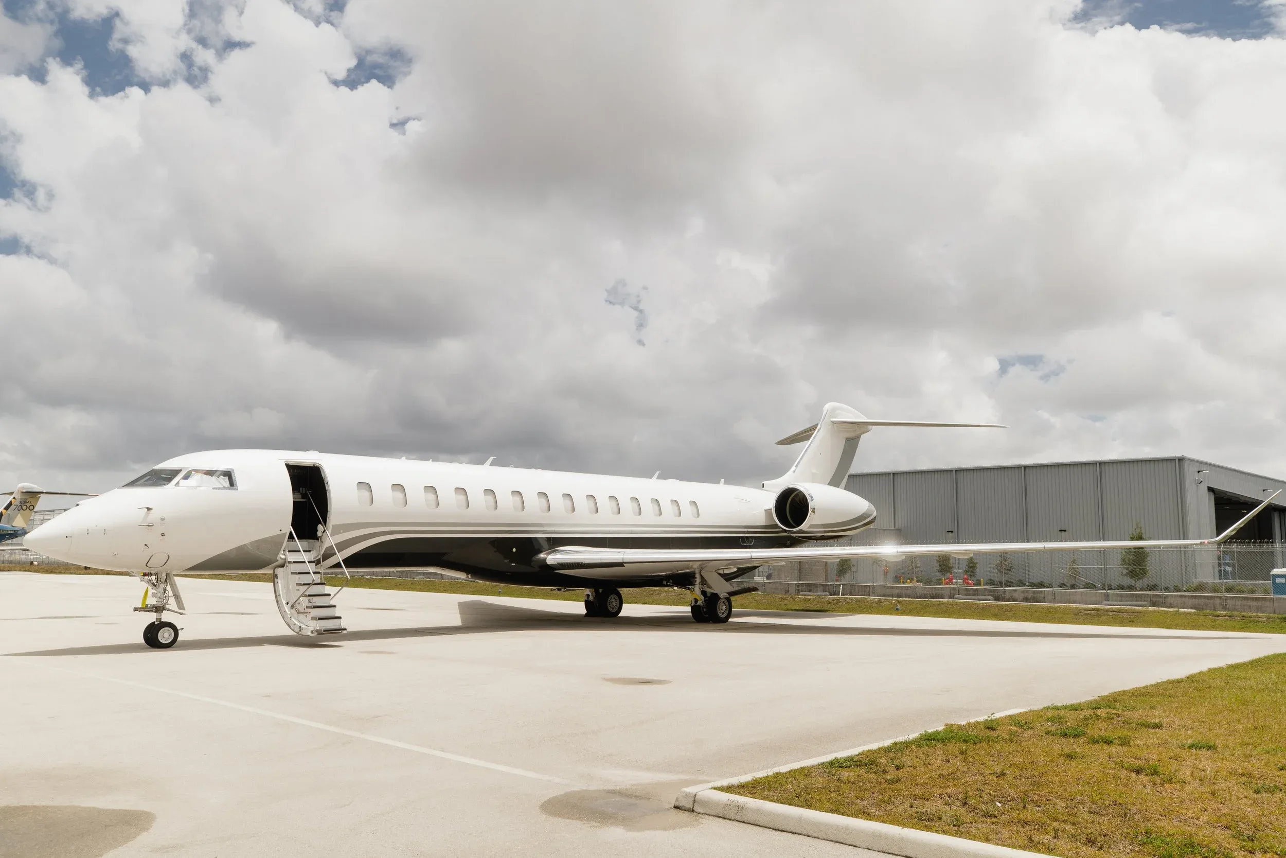 A white and grey private jet with its door open and stairs extended, parked on an airfield under a cloudy sky.