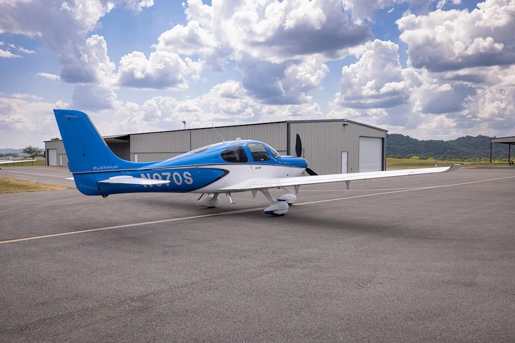 A rear-side view of the blue and white Cirrus SR22 aircraft, N970S, parked on an asphalt ramp with hangars in the background.