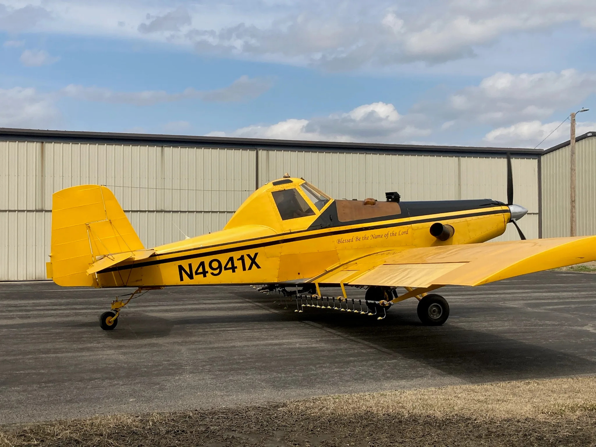 A yellow and black crop duster aircraft, N4941X, with 'Blessed be the Name of the Lord' painted on its side, parked in front of a hangar.