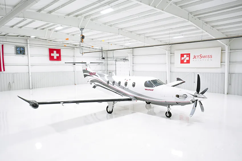 A white Pilatus PC-12 aircraft with gray and red stripes is parked in a brightly lit hangar.