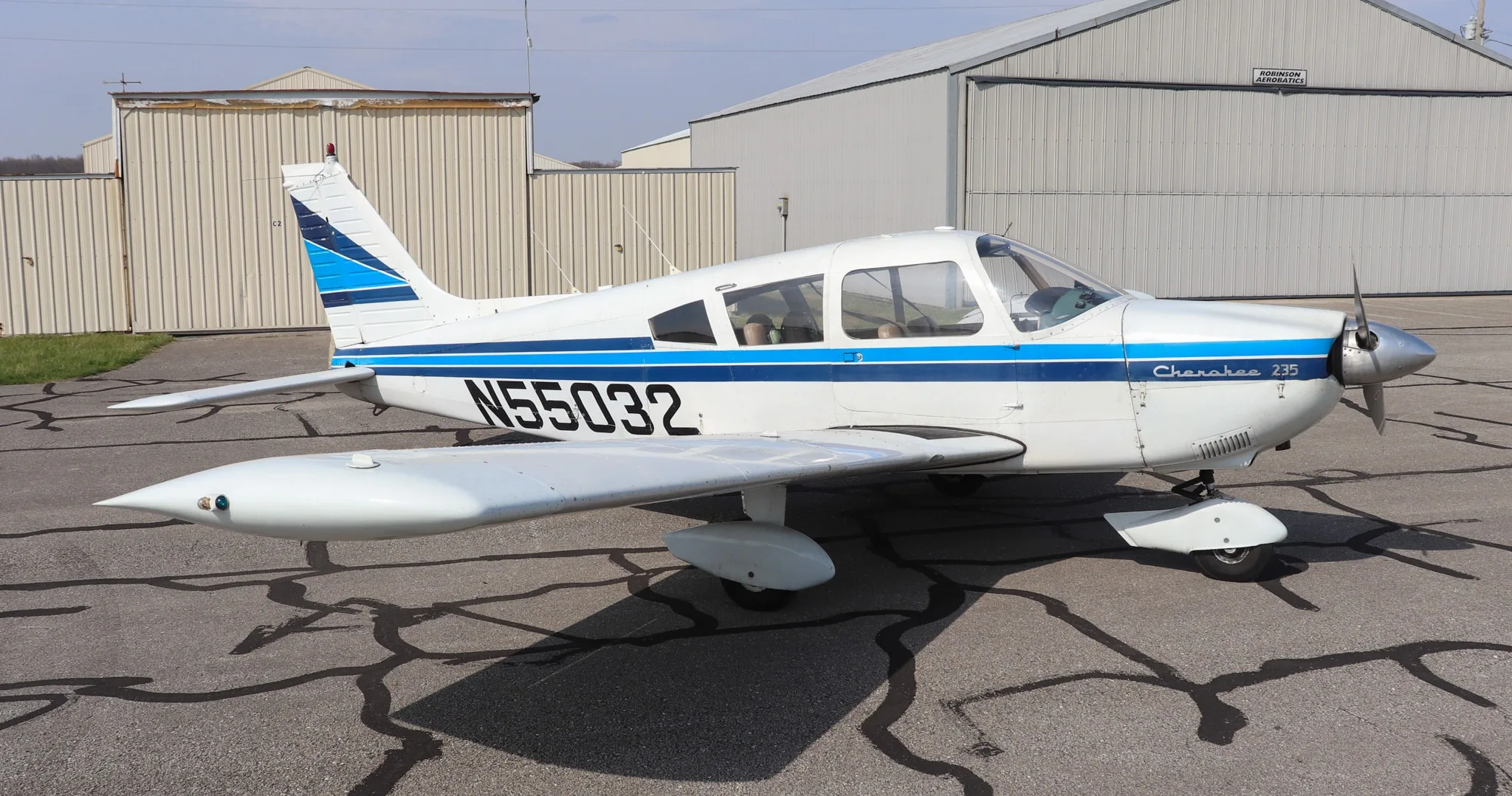 A white Piper Cherokee 235 aircraft with blue stripes and registration N55032 parked on an asphalt ramp next to hangars.