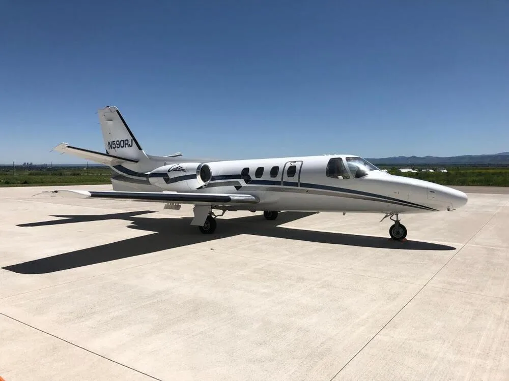 A white Cessna Citation jet with dark blue and grey stripes, registration N590RJ, parked on an airport apron under a clear blue sky.