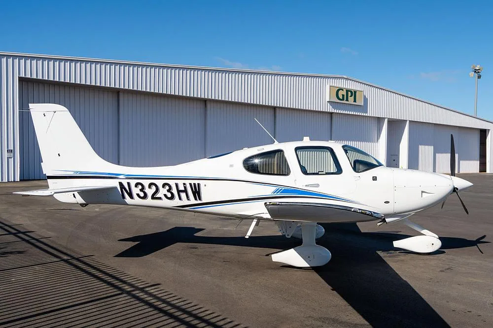 A white Cirrus SR22 aircraft with blue and black stripes, registration N323HW, parked on an asphalt ramp in front of a white hangar with a "GPI" logo.