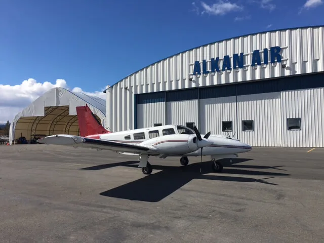 A white twin-engine propeller aircraft with a red tail and stripe is parked on an apron in front of an Alkan Air hangar under a blue sky.