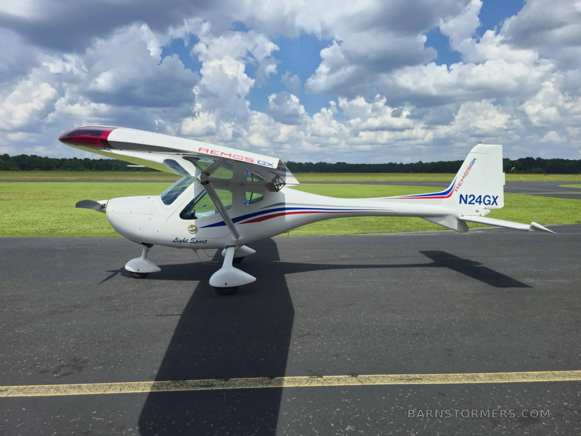 A white Remus GX Light Sport aircraft with red and blue stripes is parked on an asphalt runway under a cloudy sky.