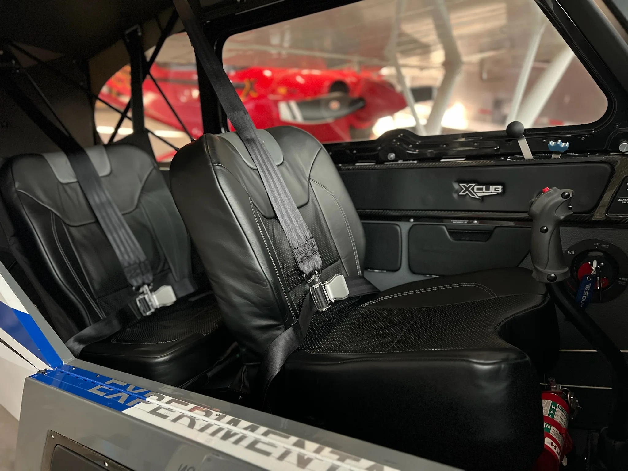 The interior cabin of an X-Cub aircraft, showing two black leather seats with seatbelts and a red car visible outside.