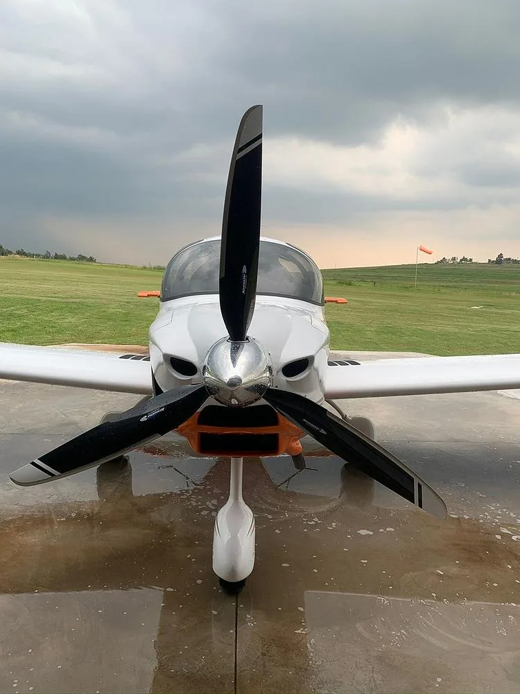 A front view of a white and orange light aircraft with a large propeller, parked on a wet surface under a cloudy sky.