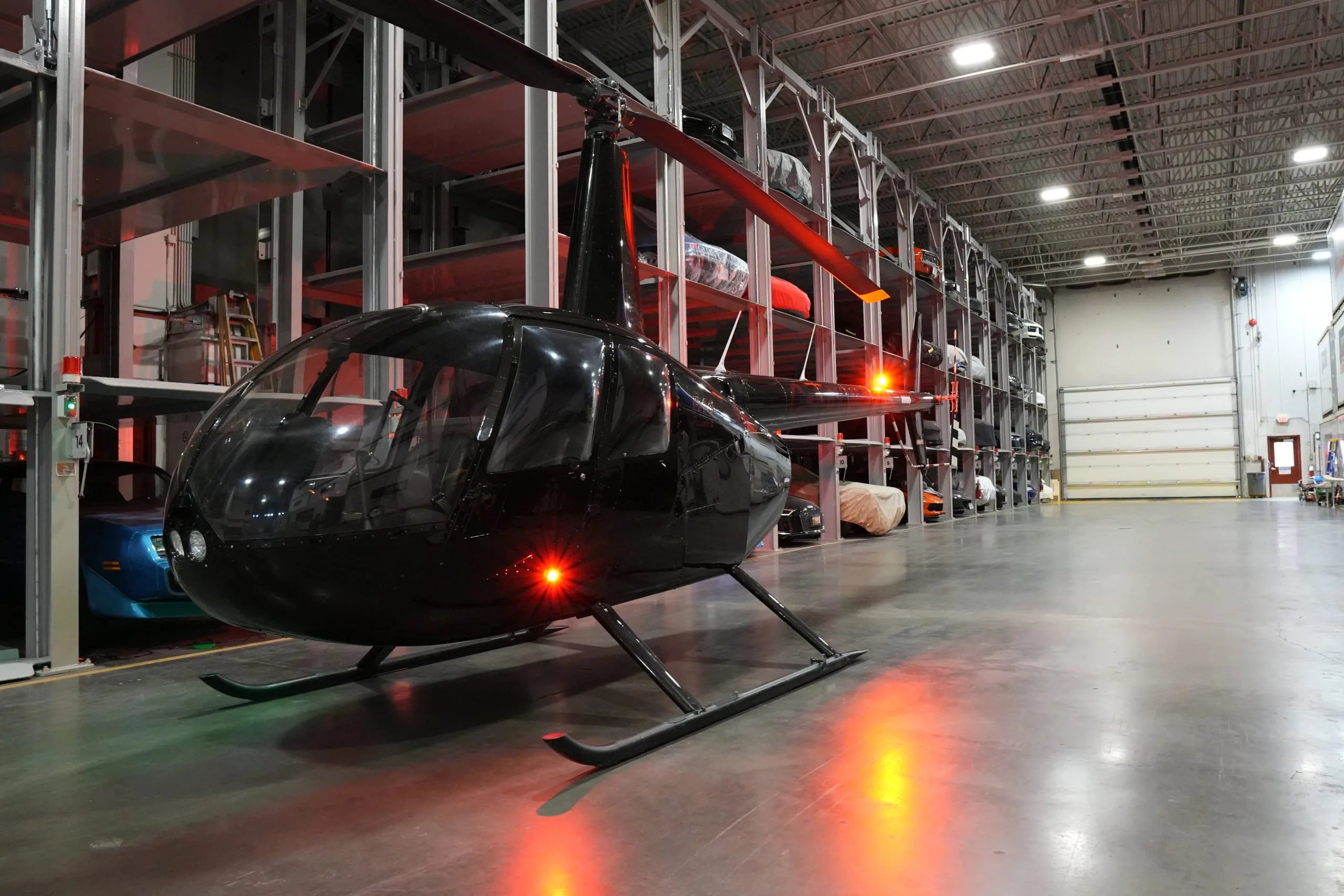 A black Robinson R44 helicopter with lights on, parked in a hangar with multi-level vehicle storage.
