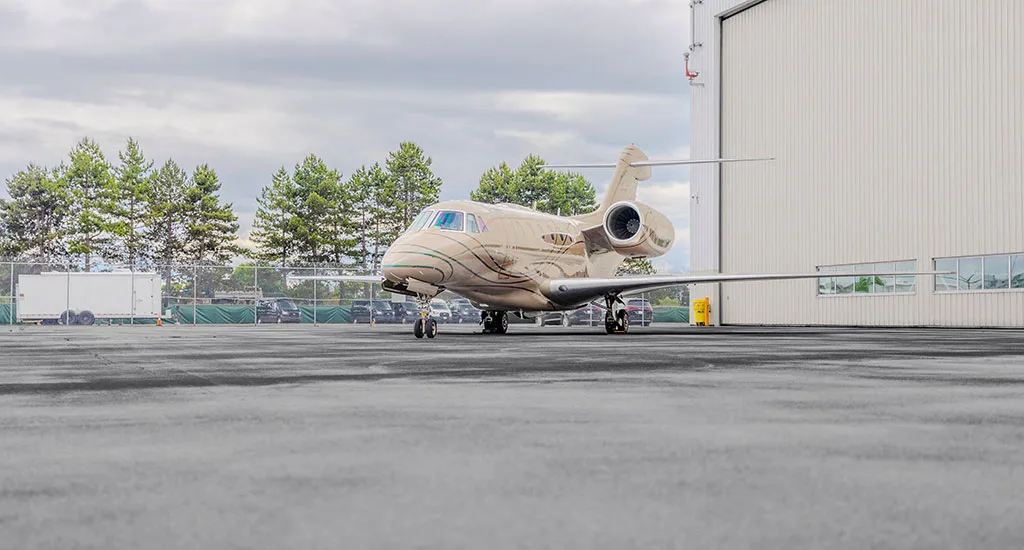 A beige business jet with green and brown stripes parked on an asphalt apron next to a white hangar under a cloudy sky.