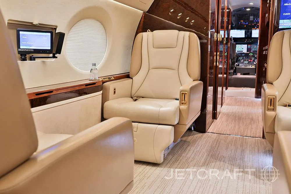A view from the private jet's cabin, showing a beige leather seat in the foreground and the cockpit visible in the distance.