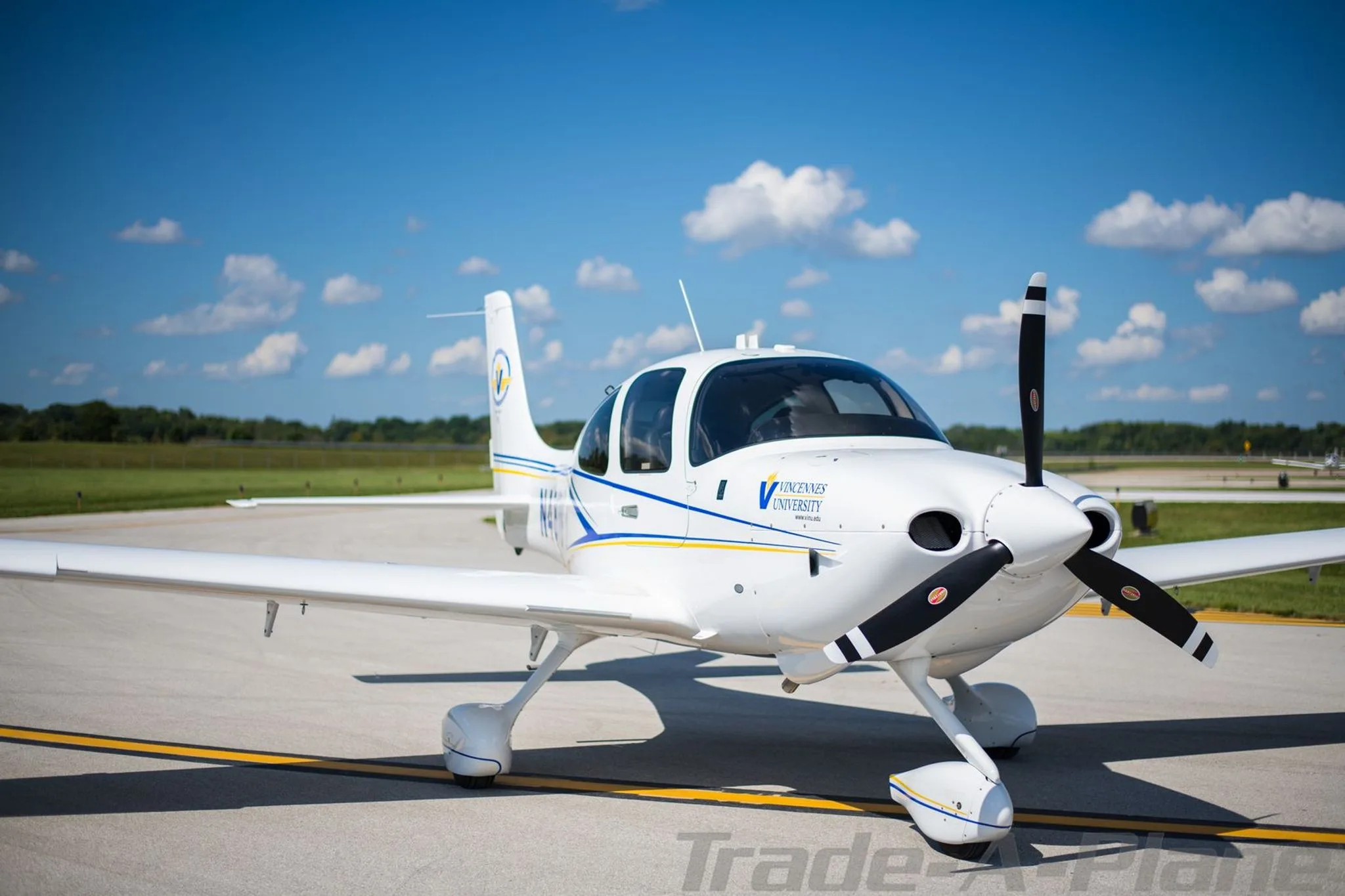 A white single-engine propeller aircraft, N42VU, with blue and yellow stripes and Vincennes University logo, parked on an airfield under a blue sky.