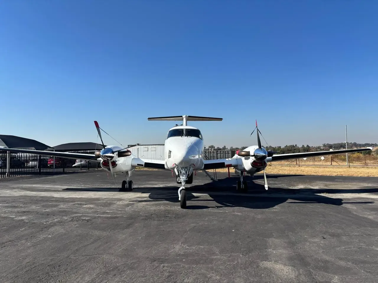 Frontal view of a white twin-engine turboprop aircraft with red accents parked on an asphalt apron under a clear blue sky.