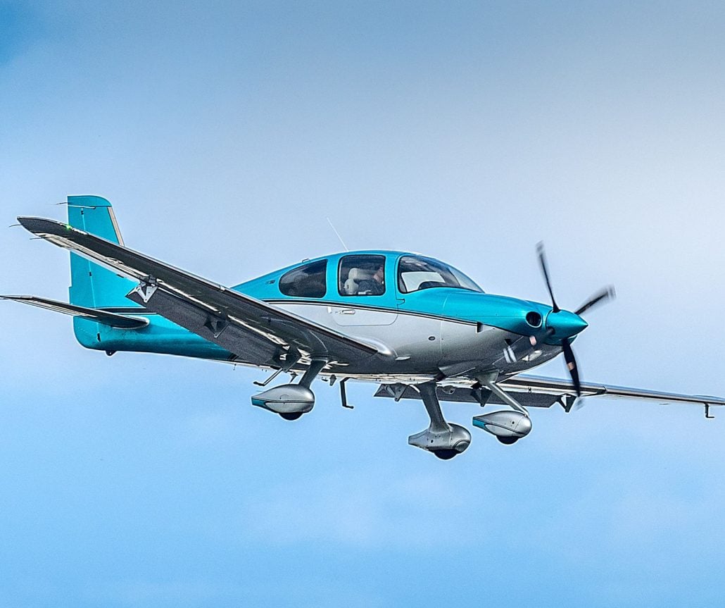 A turquoise and silver single-engine aircraft is in flight against a blue sky.