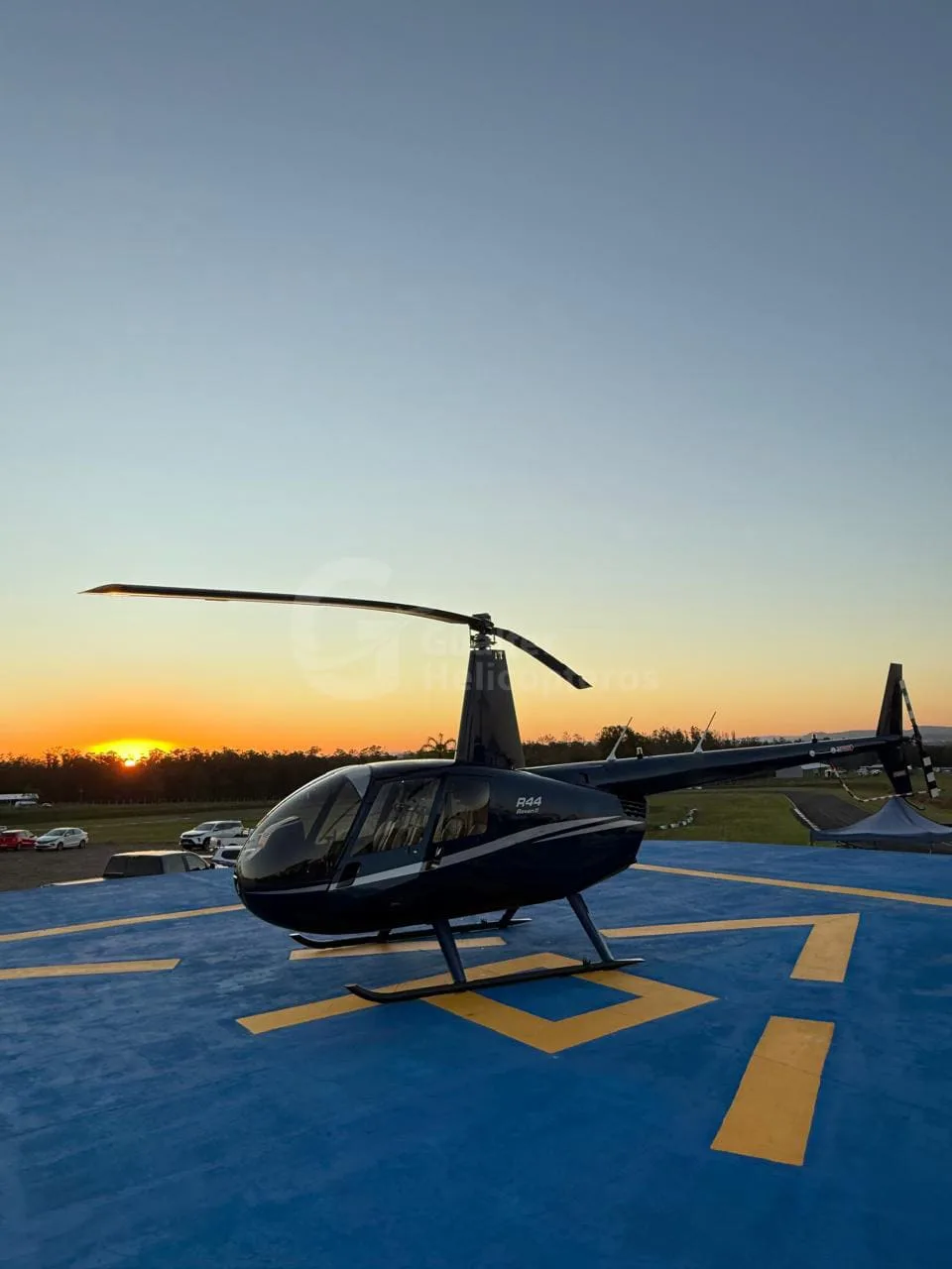 A blue Robinson R44 Raven II helicopter parked on a blue helipad at sunset.
