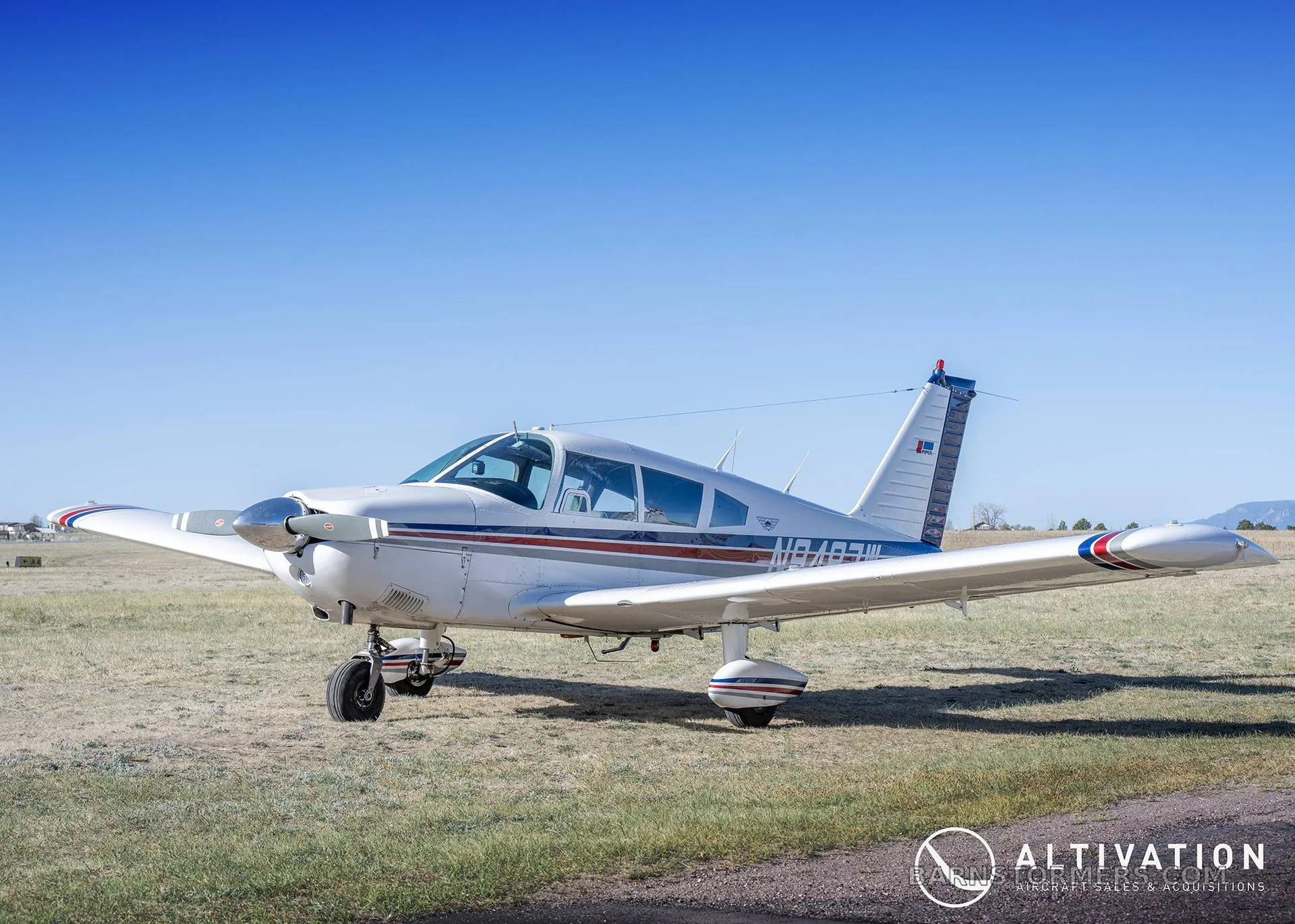 A white single-engine propeller aircraft with blue and red stripes is parked on a grassy field with distant mountains under a clear blue sky.