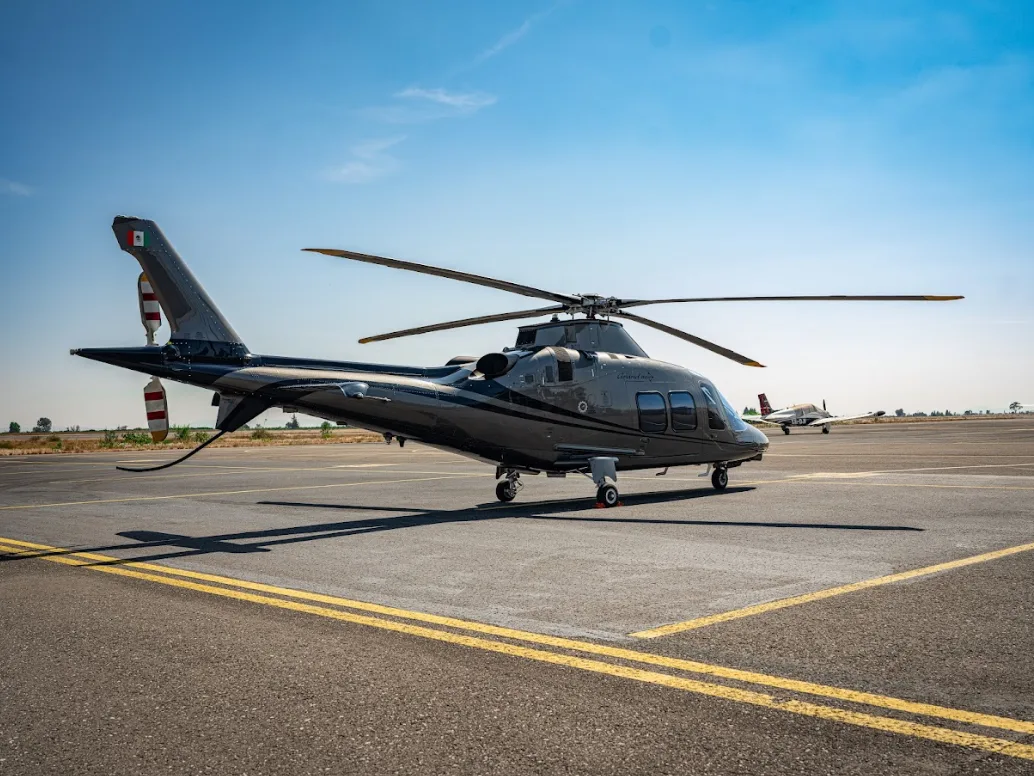 A grey AgustaWestland AW109 GrandNew helicopter is parked on an airfield with a small propeller plane in the background.