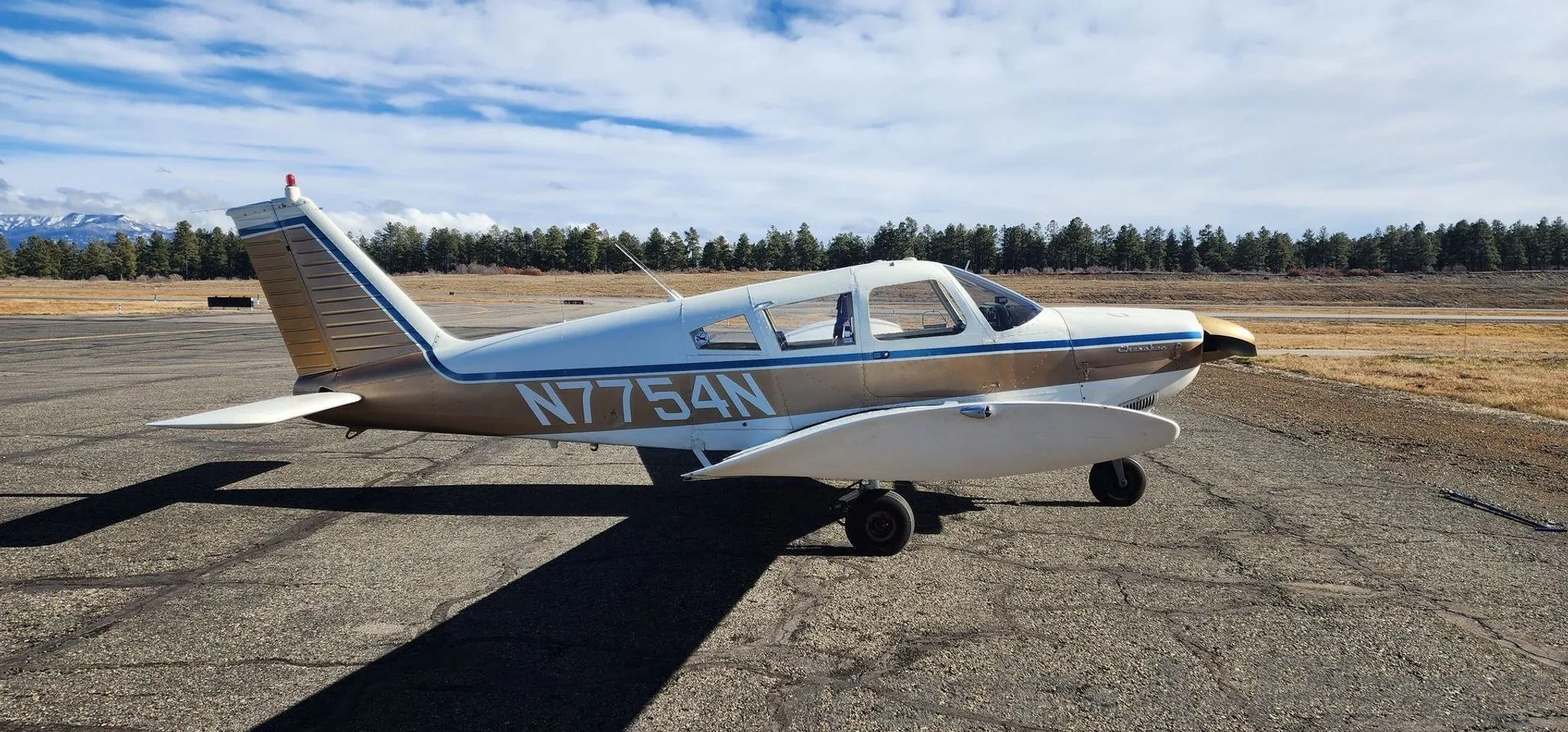 A side view of a white and gold Piper Cherokee 140 (N7754N) on an airfield, facing right.