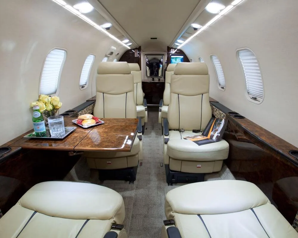 The luxurious interior cabin of a private jet with light beige leather seats, a polished wooden table, and a vase of yellow roses.