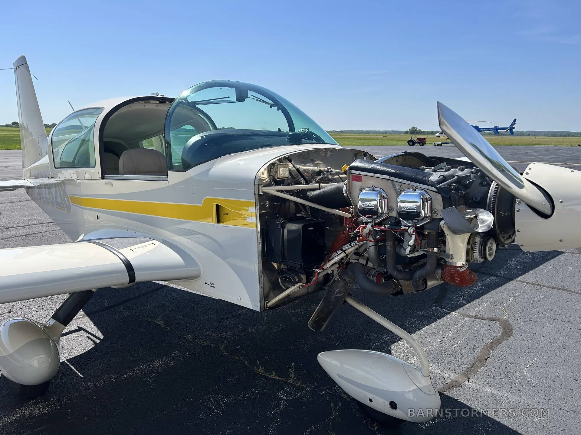 A white and yellow Grumman AA-5B Tiger aircraft with its engine cowling open, revealing the engine, parked on an airfield.