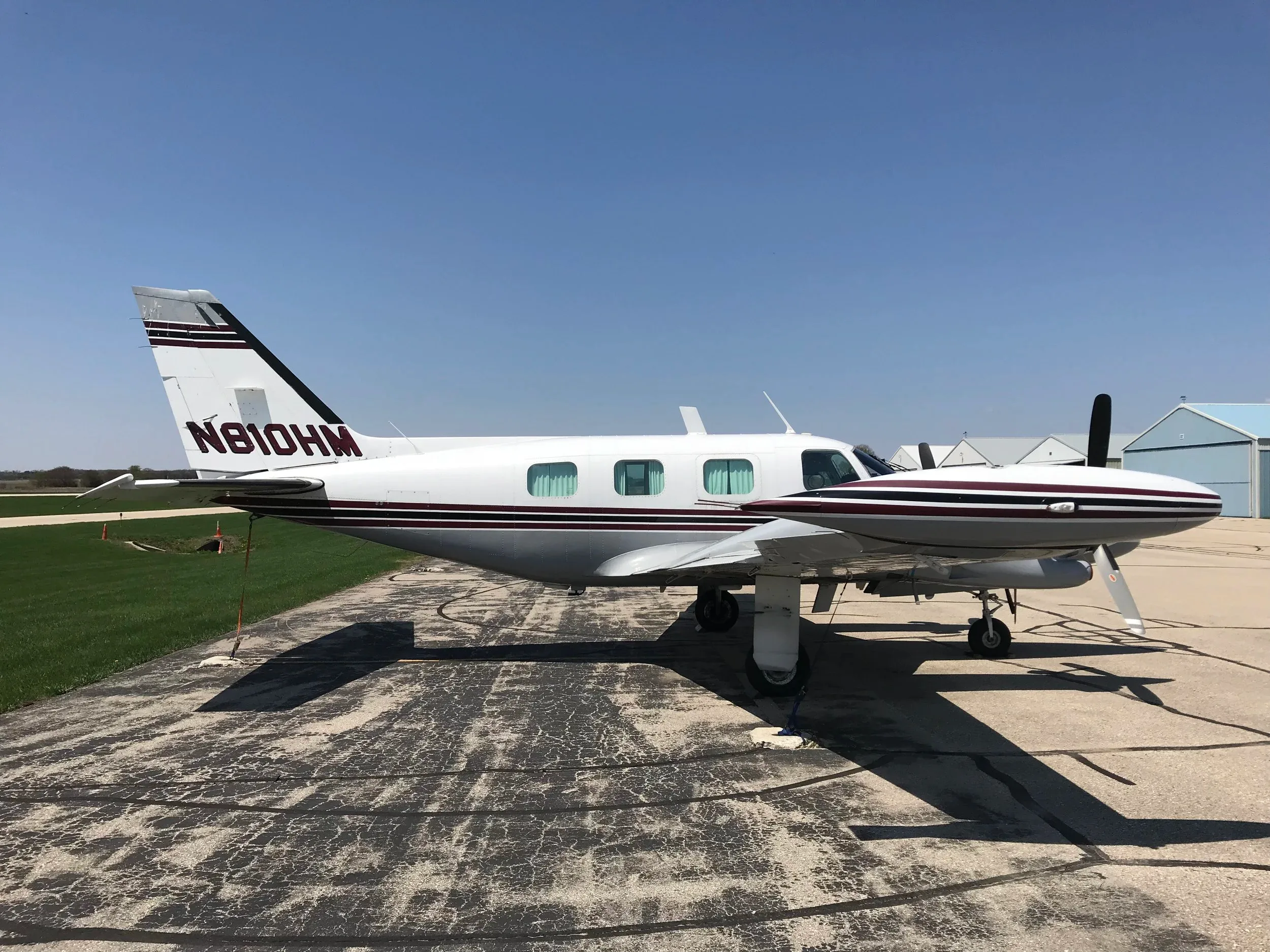 A white twin-engine propeller aircraft with maroon and black stripes, tail number N810HM, parked on an asphalt ramp under a clear blue sky.