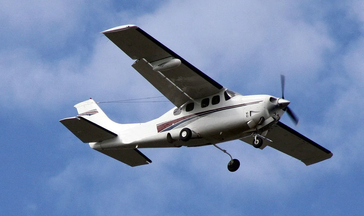 A white high-wing single-engine propeller aircraft with dark stripes in flight against a blue sky.