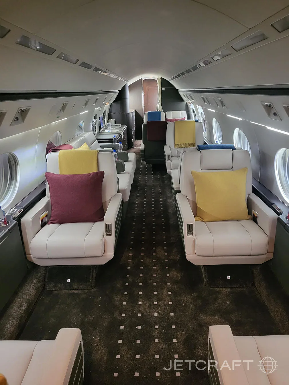 The luxurious cabin interior of a business jet with light grey seats, colorful pillows, and dark patterned carpet.